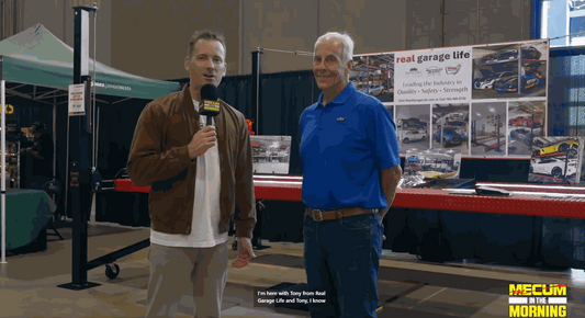Two men at Mecum in the Morning event standing near Real Garage Life booth with promotional banner in background